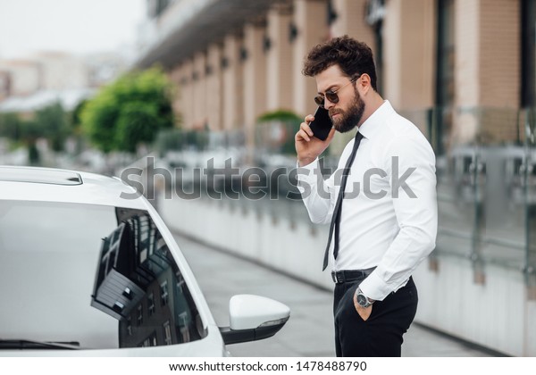 Handsome, serious, bearded manager\
in sunglasses in, speaking by phone and standing near his car\
outdoors on the streets of the city near the modern office\
center.