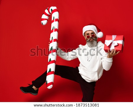 Handsome serious brutal mature bearded man in santa hat, hoodie, trendy sunglasses dancing with gift and huge candy cane. Merry Christmas and Happy New Year. Studio shot isolated on red background