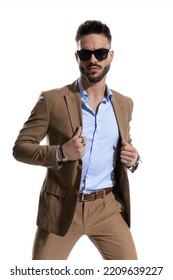 Handsome Serious Man With Open Collar Shirt Arranging Brown Suit And Posing On White Background In Studio