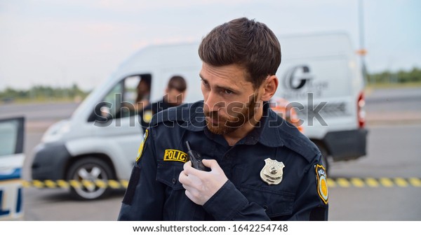 Handsome serious police officer speaking by\
walkie-talkie at road accident area. On background his colleague\
talking with a photographer on\
evidence.