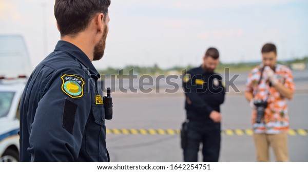 Handsome serious police officer speaking by\
walkie-talkie at road accident area. On background his colleague\
talking with a photographer on\
evidence.