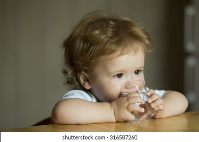 Handsome Serious Sweet Little Boy Child With Brown Hair Hazel Eyes Looking Away Sitting At Table Drinking Water From Small Glass Holding By Hands Indoor On Blur Grey Background Closeup, Horizontal