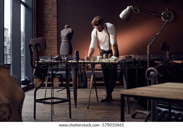 Handsome serious tailor in white shirt with
brown leather suspenders working near wooden table with threads,
apron and scissors in amazing atelier with antique furniture and
mannequin on
background