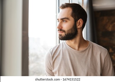 Handsome serious young bearded man standing at the window at home, looking away - Shutterstock ID 1674151522