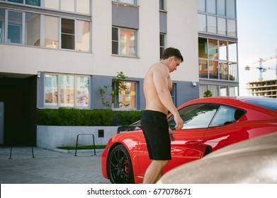 Handsome sexy male athlete man with naked torso in yard of house in parking lot Near an expensive prestigious red sports racing car, an early sunny summer morning