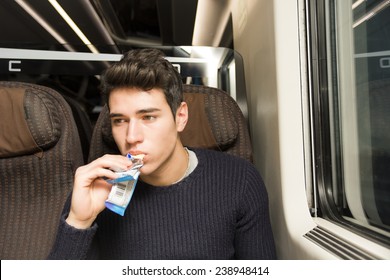 Handsome Sexy Young Man On A Train Sitting In His Passenger Seat Eating A Cereal Bar For A Healthy Snack And Looking Off To The Side With A Serious Expression