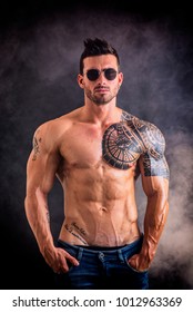 Handsome Shirtless Muscular Man With Jeans, Standing And Wearing Sunglasses, On Dark Smoky Background In Studio Shot. Tattoo Reads: We Are Free To Start Again, In Italian