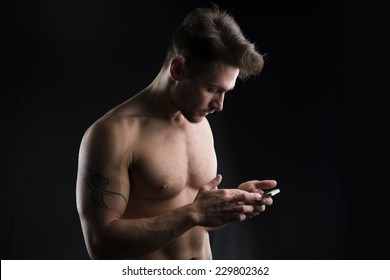 Handsome Shirtless Muscular Young Man Using Cell Phone On Dark Background