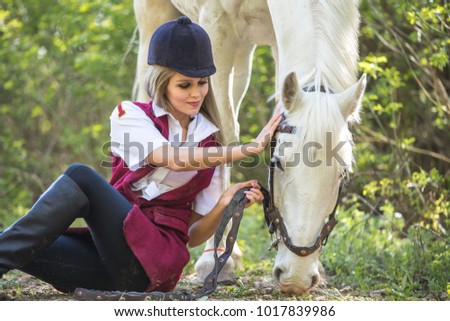 Handsome slim woman sitting on the ground with brown horse near her.