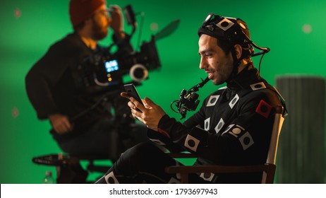 Handsome Smiling Actor Wearing Motion Capture Suit And Head Rig Having Lunch Break, Sitting On Chair, Uses Smartphone. Studio High Budget Movie. On Film Studio Period Costume Drama Film Set