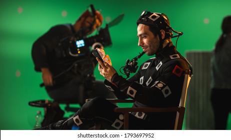 Handsome Smiling Actor Wearing Motion Capture Suit And Head Rig Having Lunch Break, Sitting On Chair, Uses Smartphone. Studio High Budget Movie. On Film Studio Period Costume Drama Film Set