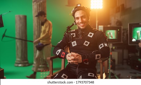 Handsome Smiling Actor Wearing Motion Capture Suit And Head Rig Having Lunch Break, Sitting On Chair, Looking At Camera. Studio High Budget Movie. On Film Studio Period Costume Drama Film Set