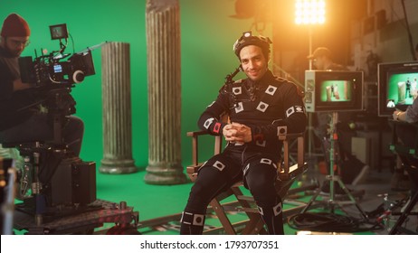 Handsome Smiling Actor Wearing Motion Capture Suit And Head Rig Having Lunch Break, Sitting On Chair, Looking At Camera. Studio High Budget Movie. On Film Studio Period Costume Drama Film Set