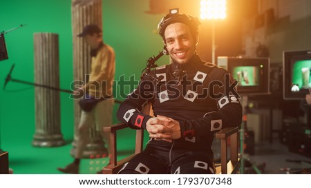 Handsome Smiling Actor Wearing Motion Capture Suit and Head Rig having Lunch Break, Sitting on Chair, Looking at Camera. Studio High Budget Movie. On Film Studio Period Costume Drama Film Set