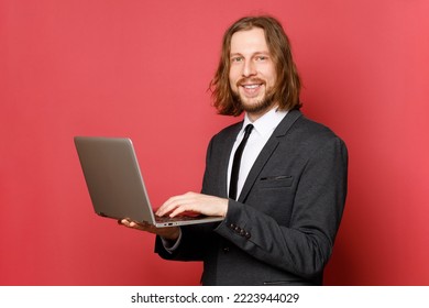 Handsome Smiling Adult Businessman With A Beard And Long Hair In A Formal Suit With A Laptop On A Red Background. 