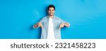 Handsome smiling adult man introduce product, pointing fingers down at promotion, standing against blue background.