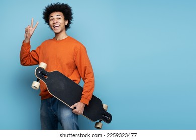 Handsome smiling African American man holding longboard showing victory sign isolated on blue background. Portrait of attractive happy skater looking at camera, copy space 