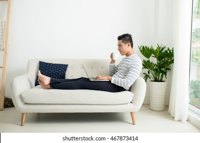 Handsome Smiling Asian Man Sitting On The Sofa And Drinking Coffee While Using Laptop At Home.