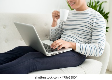 Handsome Smiling Asian Man Sitting On The Sofa And Drinking Coffee While Using Laptop At Home.