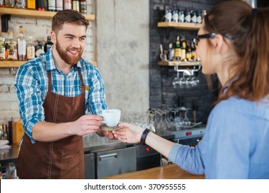 Handsome smiling barista with beard giving white cup of coffee on wooden coaster to young woman in glasses in cafe