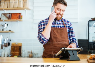 Handsome smiling barista with beard taking order on cell phone and using tablet in cafeteria - Powered by Shutterstock