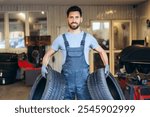 Handsome, smiling, bearded man wearing uniform, professional repairman holding tire, wheels looking at camera working in service station. Work concept