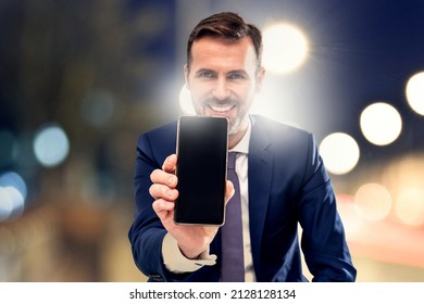 Handsome Smiling Businessman Showing Phone With Blank Black Screen To The Camera. Man Holding Smart Mobile Cell Phone In Hand. A Lot Of Copy Space.