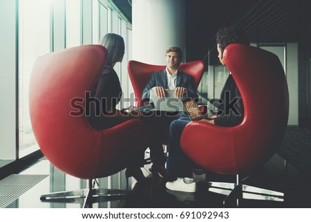 Handsome smiling businessman and two of his female colleagues are sitting on curved red armchairs with gadgets while finishing their work meeting in office interior of business skyscraper near window