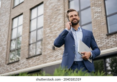 Handsome Smiling Businessman Wearing Stylish Suit Talking On Mobile Phone, Holding Laptop Standing On The Street. Successful Business	