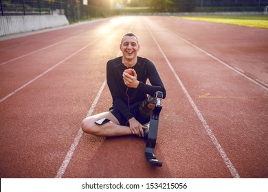 Handsome smiling caucasian sporty handicapped young man in sportswear and with artificial leg sitting on racetrack, listening music and eating apple. - Powered by Shutterstock