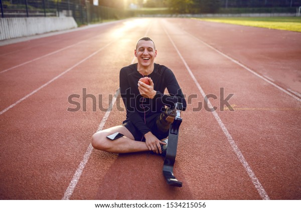 Handsome smiling caucasian sporty\
handicapped young man in sportswear and with artificial leg sitting\
on racetrack, listening music and eating\
apple.