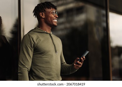 Handsome Smiling Curly Haired African Man Using Mobile Phone While Standing Outdoors, Happy Black Man With Smartphone Near Glass Building In City Looking At His Smartphone