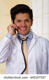 Handsome Smiling Doctor With An Stethoscope Holding From His Neck, Using His Celphone In Office Background