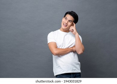 Handsome Smiling Friendly Asian Man In Casual White Plain T-shirt Thinking With Finger On Head And Looking Upward To Copy Space On Gray Isolated Background