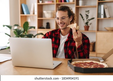 Handsome Smiling Guy Eating Pizza And Working With Laptop While Sitting At Office