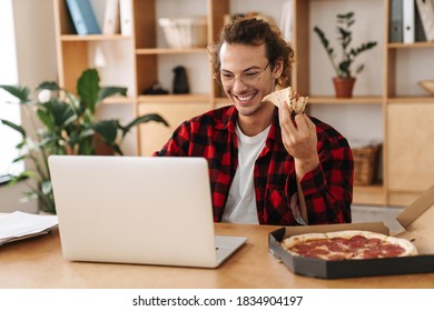 Handsome Smiling Guy Eating Pizza And Working With Laptop While Sitting At Office