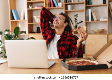 Handsome Smiling Guy Eating Pizza And Working With Laptop While Sitting At Office