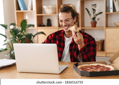 Handsome Smiling Guy Eating Pizza And Working With Laptop While Sitting At Office