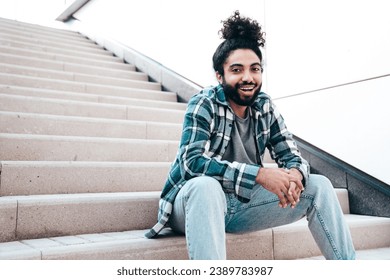 Handsome smiling hipster model. Unshaven Arabian man dressed in summer casual clothes, jeans and shirt. Fashion male with long curly hairstyle posing in the street. Sitting at the stairs - Powered by Shutterstock