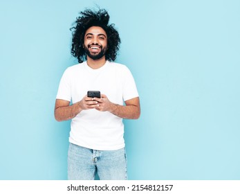 Handsome smiling hipster  model.Sexy Arabian man in summer stylish clothes.Fashion male with long curly hairstyle posing near blue wall in studio.Looking at cellphone screen. Using apps - Shutterstock ID 2154812157