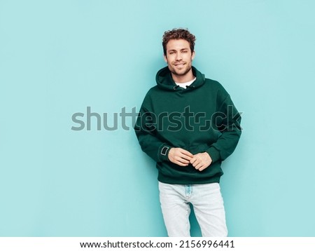 Handsome smiling hipster  model.Sexy unshaven man dressed in summer stylish green hoodie and jeans clothes. Fashion male with curly hairstyle posing in studio. Isolated on blue