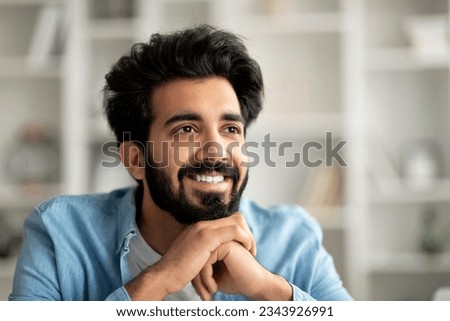 Handsome smiling indian male resting chin on hands and looking aside at copy space, happy young eastern man daydreaming in home interior, closeup portrait of happy bearded millennial guy