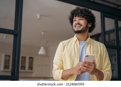 Handsome Smiling Indian Man Holding Mobile Phone Shopping Online Looking Away At Home, Copy Space 