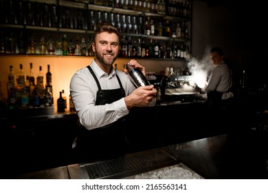 Handsome Smiling Male Bartender In White Shirt And Black Apron Holding Steel Shaker In Hands. Blurred Bottles On Shelves In The Background