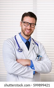 Handsome Smiling Male Doctor Portrait With Glasses, Arms Crossed And Stethoscope On His Neck Standing Close Against White Window Blinds