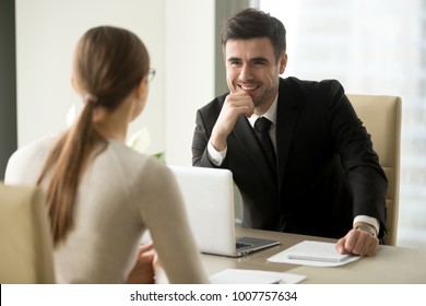 Handsome Smiling Male Office Worker In Business Suit Sitting At Desk With Laptop In Front Of Female Colleague. Happy Businessman Talking With Secretary. Positive First Impression On Job Interview