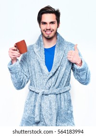 Handsome Smiling Man Or Bearded Sexy Guy In Blue Terry Bathrobe Or Dressing Gown Holds Coffee Or Tea Cup Shows Thumb Up Isolated On White Background