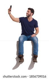 Handsome Smiling Man In Boots, Jeans And Unbuttoned Lumberjack Shirt Is Sitting On A Top, Holding Telephone And Taking A Selfie. Full Length Studio Shot Isolated On White.