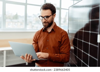 Handsome smiling man, engineer, programmer holding laptop computer planning startup working with solar panel in modern office. Innovation, technology,  solar energy concept