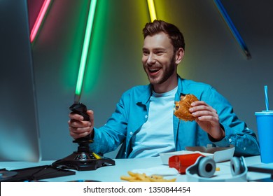 Handsome And Smiling Man Holding Tasty Burger And Playing Video Game With Joystick